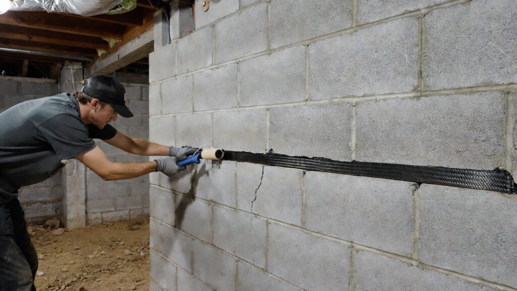 A homeowner repairing a horizontal foundation crack in a basement wall using a carbon fiber strap and structural epoxy. A homeowner repairing a horizontal foundation crack in a basement wall using a carbon fiber strap and structural epoxy.