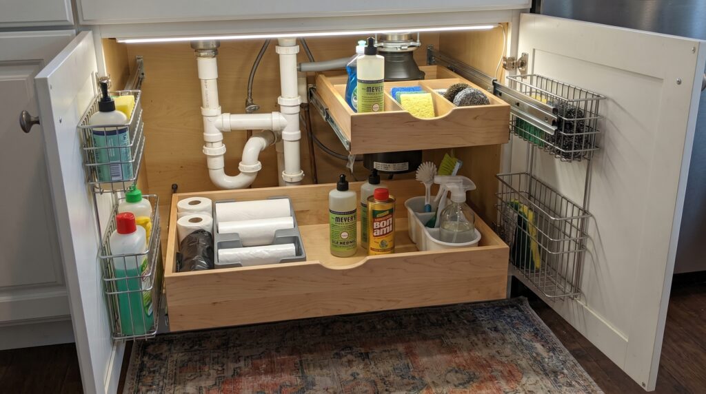 Under sink pull out drawers installed below a kitchen sink with cleaning supplies organized around plumbing pipes