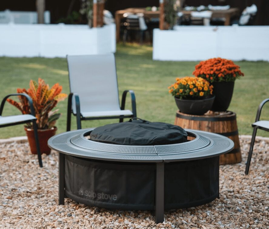 Solo Stove Surround Fire Pit Table and Barrier A burning Solo Stove fire pit placed securely inside the Surround protective barrier on a backyard patio at dusk. Two drinks and a bowl of snacks rest safely on the perforated metal tabletop, demonstrating the cool-to-touch safety feature.