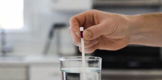Water Hardness Test Strips: How to Read & Use Them (2026) A person dipping a water hardness test strip into a glass of tap water to check for hard water.
