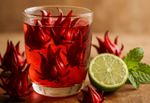 Hibiscus Tea Benefits: Roselle Science, Brewing, Safety & Growing Guide Glass of bright red hibiscus tea with roselle petals floating inside, lime half and fresh mint on a wooden surface nearby.